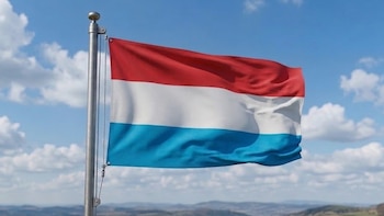 Bandera de Luxemburgo ondeando en un mástil metálico. Presenta tres franjas horizontales: roja, blanca y azul claro. Fondo de cielo azul con nubes y un paisaje rural con colinas y un pueblo