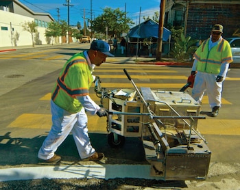 Trabajador en chaleco reflectante opera máquina para pintar una línea blanca en un paso de cebra amarillo en Los Ángeles. Otro trabajador con sopladora cerca