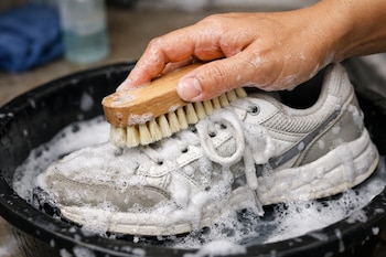 Una mano cepilla un tenis blanco cubierto de espuma de jabón dentro de un recipiente negro, con agua jabonosa y un cepillo de madera visible.