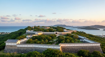 Castillo de Nakagusuku con muros de piedra tradicional, edificaciones blancas y techos oscuros, rodeado de vegetación verde, mar y cielo con nubes.