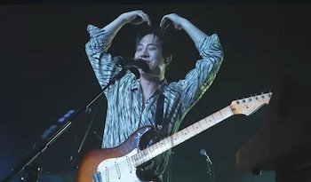Hombre sonriente con camisa de rayas tipo cebra y una guitarra eléctrica en un escenario oscuro, sus manos levantadas a la cabeza mientras canta en un micrófono