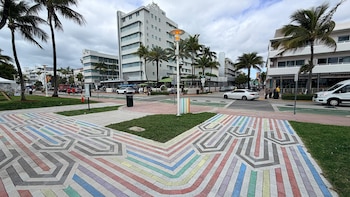 Vista de un paseo peatonal con patrón de ladrillos de colores en Lummus Park, Miami Beach, con palmeras, edificios y personas en segundo plano
