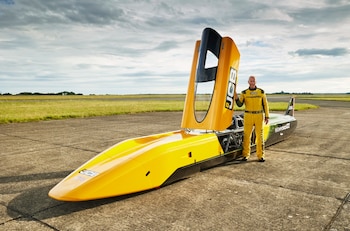 Hombre con uniforme de piloto amarillo de pie junto a un vehículo aerodinámico amarillo y negro sobre asfalto, con campo verde y cielo nublado de fondo