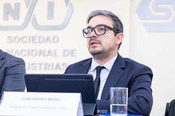 Un hombre con anteojos y traje formal sentado frente a un letrero con su nombre y un vaso de agua, ante un panel corporativo