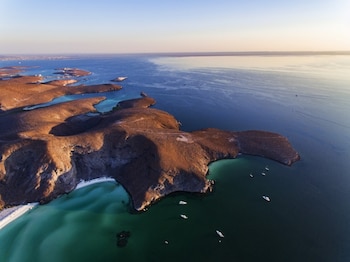 Vista aérea de una costa desértica con colinas marrones, playas de arena blanca, agua turquesa y azul, y varias embarcaciones