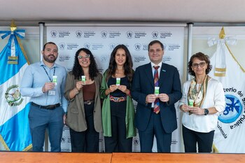 Cinco personas de pie, sonrientes, sostienen pequeños empaques verdes y blancos frente a un banner del Ministerio de Salud Pública de Guatemala y banderas