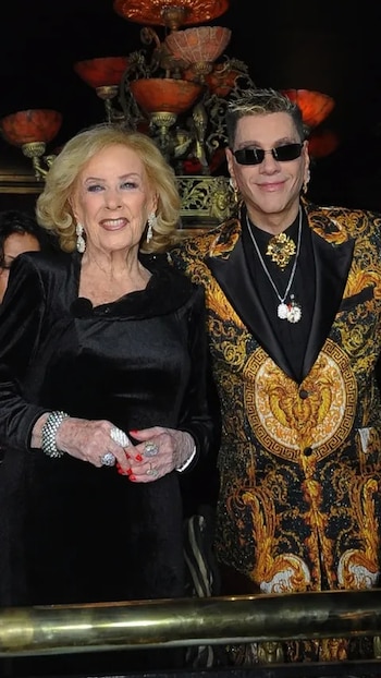 Mirtha Legrand, con cabello rubio y vestido negro, posa sonriente junto a Roberto Piazza, quien usa un traje barroco dorado y negro con gafas de sol oscuras