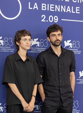 Una mujer con gafas y un hombre con barba vestidos de negro posan frente a un fondo azul con letras y logotipos blancos