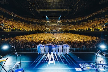 Vista trasera de Jorge Drexler y su banda inclinándose en el escenario, frente a una multitud masiva y aplaudiendo en el Movistar Arena iluminado