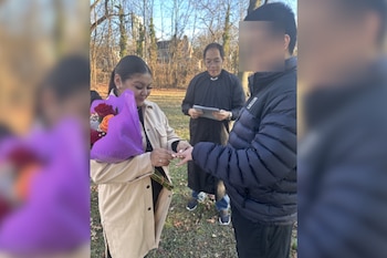 Una mujer con un ramo de flores púrpura coloca un anillo en el dedo de un hombre frente a un oficiante con una tablet en un entorno exterior con árboles