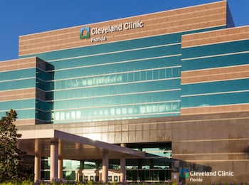 Exterior del edificio Cleveland Clinic Florida con fachada de vidrio y piedra. Se ven columnas, entrada principal, y jardines con flores