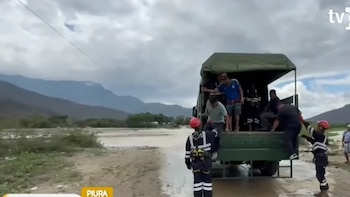 Personas descienden de la tolva de un camión militar con el apoyo de rescatistas con cascos junto a un río crecido y colinas bajo cielo nublado