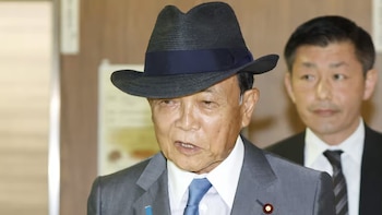 Un hombre de edad avanzada con sombrero oscuro, traje gris y corbata azul, junto a otra persona de traje detrás