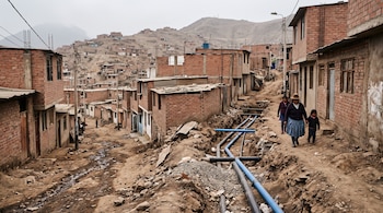 Calle de tierra con tuberías en una zanja, casas de ladrillo sin tarrajear y tres personas caminando.