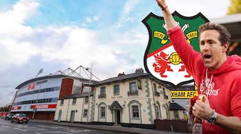 Ryan Reynolds con una sudadera roja y el brazo levantado junto al escudo del Wrexham AFC, un pub y un estadio de fútbol.