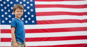 Niño de pie sonriente frente a una bandera grande de Estados Unidos. Viste una camiseta azul y pantalones cortos. La bandera muestra estrellas y rayas rojas y blancas.