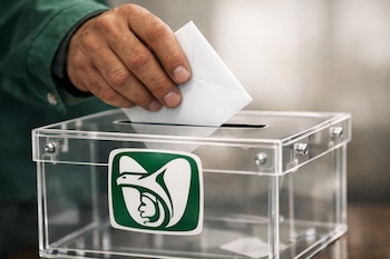 Mano de persona con manga verde deposita un sobre blanco en una urna de acrílico transparente con el logo del IMSS de un águila estilizada en color verde.