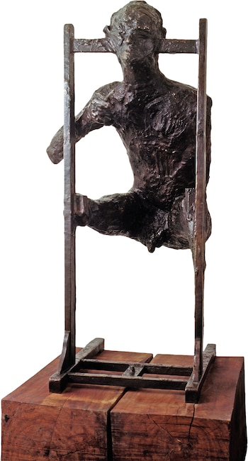 Escultura de bronce de una figura humana incompleta apoyada en una estructura vertical de metal sobre una base de madera con fondo blanco