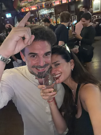 Un hombre con barba y una mujer sonrientes posan en el interior de un bar mientras ella sostiene un vaso de cristal