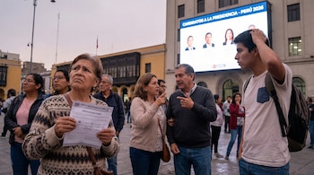 Grupo de personas de diversas edades en una plaza pública en Perú, algunas con cédulas electorales, observando una gran pantalla luminosa que muestra "Candidatos a la Presidencia - Perú 2026".