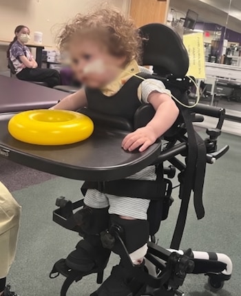 Un niño pequeño con el rostro difuminado permanece sujeto en una silla terapéutica con bandeja, donde reposa un aro de plástico amarillo