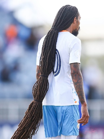 Un futbolista de espaldas lleva camiseta blanca, pantalón corto azul y rastas muy largas atadas en tramos hasta las piernas