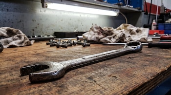 Una llave combinada de metal sobre una mesa de madera. Se ven tornillos, trapos sucios y una luz fluorescente en la parte superior.