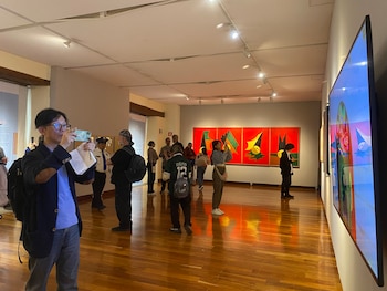 Varias personas en una sala de exposición de arte, observando cuadros de fondo rojo y figuras abstractas, y una pantalla digital. Hay piso de madera y luces en el techo