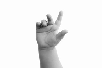 Mano de bebé en blanco y negro, con los dedos ligeramente abiertos y levantados, sobre un fondo blanco liso. Se observan los detalles de la piel infantil.