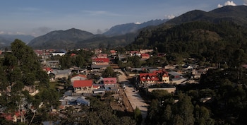Vista aérea de un pueblo en un valle, con casas de tejados rojos y naranjas, rodeado de densa vegetación y montañas boscosas bajo un cielo azul claro