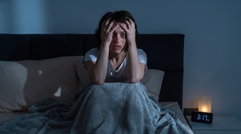 Una mujer sentada en una cama con las manos en la cabeza junto a una mesa de noche con un reloj digital y una lámpara encendida.