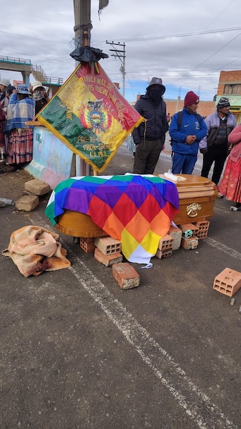 Ataúd de madera con bandera Wiphala, sobre ladrillos en calle. Personas alrededor, bandera boliviana y manta con manchas oscuras en el suelo