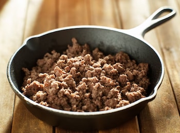 Una sartén de hierro fundido llena de carne de res molida cocida, de color marrón claro, reposa sobre una superficie de madera clara