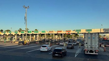 Múltiples carriles y cabinas de un peaje con vehículos en tránsito. Se observa un camión, autos y un mástil con una bandera bajo un cielo azul despejado