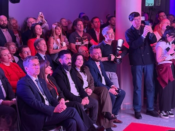 Un grupo de personas, hombres y mujeres, sentados y de pie, en un evento interior con iluminación púrpura, varios sosteniendo teléfonos y cámaras