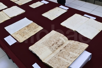 Una mesa cubierta con tela roja muestra varios documentos antiguos abiertos y cerrados con escritura manuscrita y un libro abierto, junto a tarjetas blancas