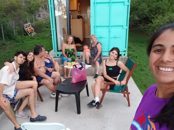 Grupo de personas sonriendo al aire libre frente a una mini casa contenedor turquesa. Incluye a una mujer mayor, niños y adultos jóvenes