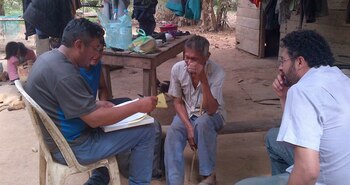 Cuatro hombres conversan al aire libre frente a una casa de madera mientras uno de ellos escribe en una carpeta
