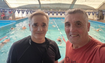 Dos hombres posan en primer plano frente a una piscina cubierta donde nadan varias personas