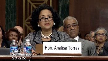 Una mujer con gafas y collar de perlas sentada frente a un letrero con el nombre Analisa Torres, botellas de agua y un micrófono