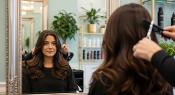Una mujer joven con cabello largo y ondulado se sienta frente a un espejo; un estilista le peina y corta el cabello con peine y tijeras.