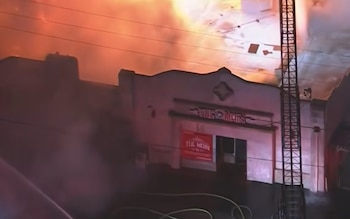 Fachada de un edificio con la inscripción The Main, humo y fuego en la parte superior, mangueras en el suelo y una escalera de bomberos
