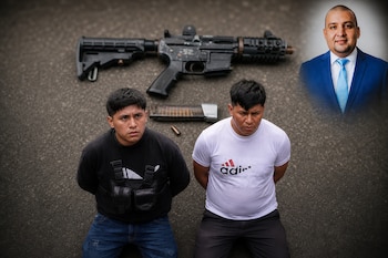 Dos hombres arrodillados sobre el suelo frente a un fusil y un cargador, acompañados por el retrato de un hombre con traje azul en una esquina.