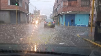 Vista desde un automóvil de una calle inundada con dos vehículos, edificios de ladrillo, un semáforo en verde y una persona en la acera.