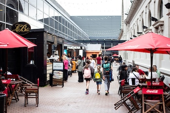 Vista de una calle con personas caminando y sentadas en mesas de café al aire libre bajo sombrillas rojas, flanqueada por edificios y locales comerciales