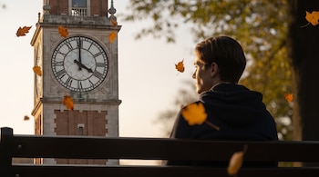 Hombre joven sentado en un banco, mirando una torre del reloj de ladrillo y piedra con esfera blanca y números romanos. Hojas de otoño flotan en el aire.