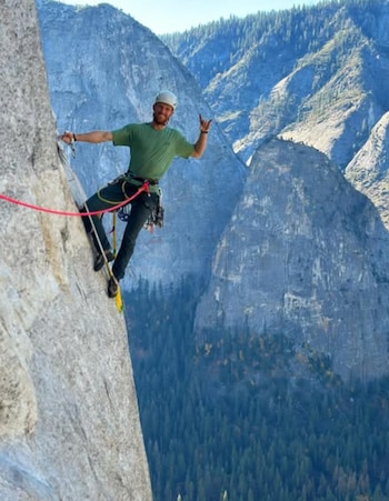Un hombre con casco blanco, camiseta verde y arnés se sujeta a una pared de roca con una cuerda roja. Al fondo se ven montañas y árboles