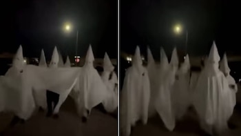 Varias personas cubiertas con sábanas y capuchas cónicas blancas caminan de noche bajo la luz de una farola