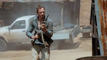 Michael Fassbender, com sangue na testa e rosto sujo, segurando uma arma e olhando para a direita. Ele está vestindo uma camisa azul desabotoada e calça clara, com uma caminhonete branca atrás dele.
