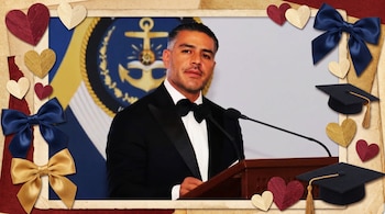 Hombre con traje y corbatín negro en podio con micrófono. Marco decorado con papel, corazones rojos, dorados, crema, moños azules, dorados, y birretes negros de graduación.